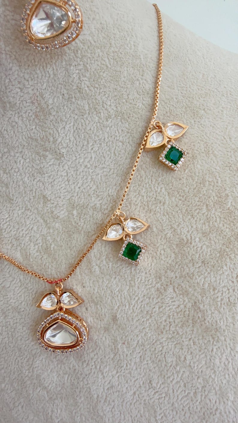 May include: A gold chain necklace with three pendants. Each pendant features a green gemstone surrounded by clear stones and two teardrop-shaped clear stones. The necklace is on a white background.
