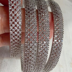 Op de afbeelding: Een set zilverkleurige armbanden bezet met sprankelende, heldere edelstenen. De ronde armbanden lijken van metaal te zijn, met een doorlopend patroon van kleine, gelijkmatig verdeelde stenen. De sieraden worden gepresenteerd tegen een lichte achtergrond.