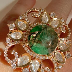 May include: A gold ring with a large green emerald gemstone surrounded by smaller clear gemstones. The ring has a delicate, ornate design.