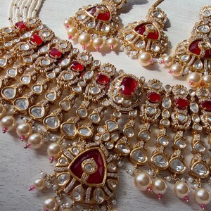 Puede incluir: Un conjunto de joyas dorado que incluye un collar y pendientes a juego. El collar y los pendientes est&aacute;n adornados con piedras preciosas rojas, cristales transparentes y detalles colgantes en forma de l&aacute;grima. Tambi&eacute;n se incorporan perlas de color crema.