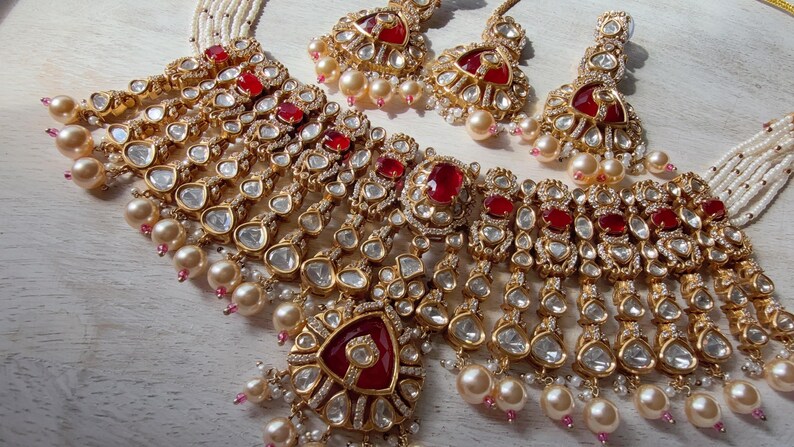 Puede incluir: Un conjunto de joyas dorado que incluye un collar y pendientes a juego. El collar y los pendientes est&aacute;n adornados con piedras preciosas rojas, cristales transparentes y detalles colgantes en forma de l&aacute;grima. Tambi&eacute;n se incorporan perlas de color crema.