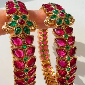 Op de afbeelding: Twee goudkleurige armbanden met een bloemmotief. Elke armband is versierd met rode en groene edelstenen. De edelstenen zijn gerangschikt in een bladvormig patroon. De armbanden worden tegen een witte achtergrond getoond.