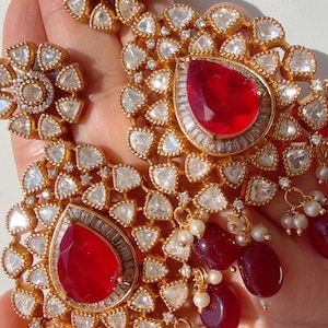 May include: Close-up of gold-toned earrings and a ring set with clear and red gemstones. The earrings feature large, teardrop-shaped red stones surrounded by clear stones and smaller red beads. The ring has a floral design.