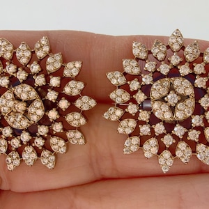 May include: A pair of gold-tone earrings with a circular design featuring a cluster of small, clear rhinestones. The earrings have a floral-like pattern.