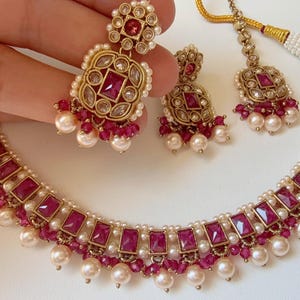 Puede incluir: Un conjunto de joyas de tono dorado con collar y pendientes. El collar y los pendientes están adornados con piedras rectangulares de color rubí, pequeñas cuentas de rubí y perlas blancas. El conjunto se muestra sobre una superficie blanca.