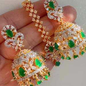 May include: A pair of gold earrings with green and white stones. The earrings are in the shape of a jhumka, a traditional Indian style of earring.