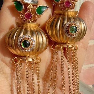 May include: A pair of ornate gold earrings. Each earring features a large, ribbed, gold-colored bulbous shape with a central green stone surrounded by small clear stones. Above the bulb is a design with clear and green stones. Gold chains hang from the bottom.