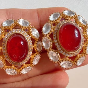 Peut inclure: Une paire de boucles d'oreilles dorées. Chaque boucle d'oreille présente une grande pierre rouge ovale au centre, entourée de pierres claires taillées en ovale. Les boucles d'oreilles ont un design circulaire avec une bordure de pierres claires plus petites. Bijou remarquable.