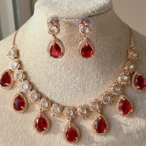 May include: A rose gold necklace and earring set with teardrop-shaped, deep red gemstones. The necklace and earrings are adorned with clear, round accent stones. The jewelry is displayed on a mannequin.