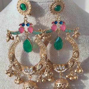 Op de afbeelding: Gouden oorringen met ingewikkelde details. Elke oorring heeft een groene edelsteen, roze en blauwe accenten en een druppelvormige groene edelsteen. Kleine gouden belletjes hangen aan de onderkant, wat het ontwerp verfraait.