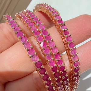 May include: Four rose gold bangles are displayed, each adorned with a row of vibrant, round, pink gemstones. The gemstones are closely set, creating a continuous band of color. The bangles are held in a hand, showcasing their design.