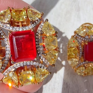 Może przedstawiać: Zestaw ozdobnej biżuterii, składający się z dużego pierścionka i pasującego kolczyka. Elementy mają złote oprawy, centralny czerwony kamień szlachetny, otoczony żółtymi i przezroczystymi kamieniami.