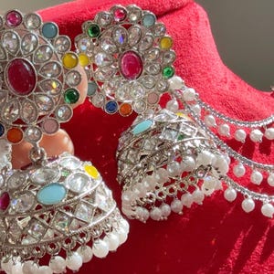 Puede incluir: Pendientes plateados con diseño floral, con una piedra roja central rodeada de gemas de colores. Los pendientes tienen forma de campana colgante con detalles de perlas blancas y una cadena de perlas.