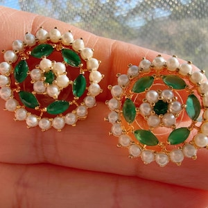 Può includere: Un paio di orecchini a bottone dorati. Ogni orecchino presenta un gruppo centrale di perle bianche e pietre preziose verdi, circondato da un anello di perle bianche. Gli orecchini hanno un design circolare.
