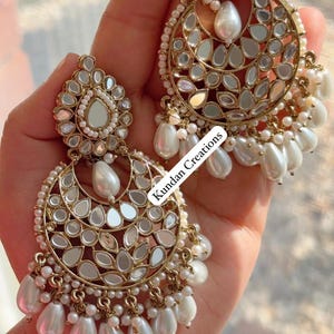 May include: Gold-toned, ornate earrings with a crescent moon design, featuring mirrored accents and dangling white pearl beads. The earrings have a floral-like top and the text "Kundan Creations".