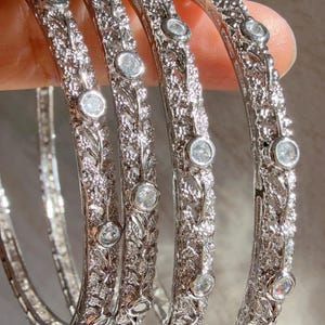 May include: Four silver bangles with detailed floral and leaf patterns. Each bangle is set with several clear, round gemstones. The bracelets are held in a hand, highlighting their intricate design and sparkling appearance.