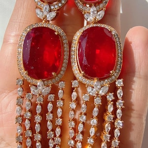 Peut inclure: Boucles d'oreilles pendantes dorées avec de grandes pierres rouges ovales et un halo de pierres claires. Les boucles d'oreilles ont une frange en cascade de pierres claires et étincelantes. Design luxueux.