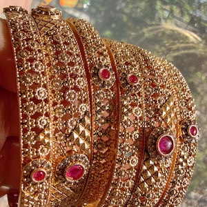 May include: A collection of gold-colored bangles with intricate floral and geometric patterns. Each bangle is adorned with small, round, red gemstones. The bangles are stacked together, showcasing their detailed design and craftsmanship.