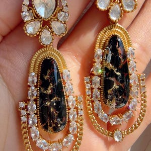 May include: Gold-toned drop earrings with a black, marbled center stone and clear, sparkling accents. The earrings feature a chain-like design and are set with multiple clear crystals. The top of the earrings have a large, clear stone surrounded by smaller crystals.