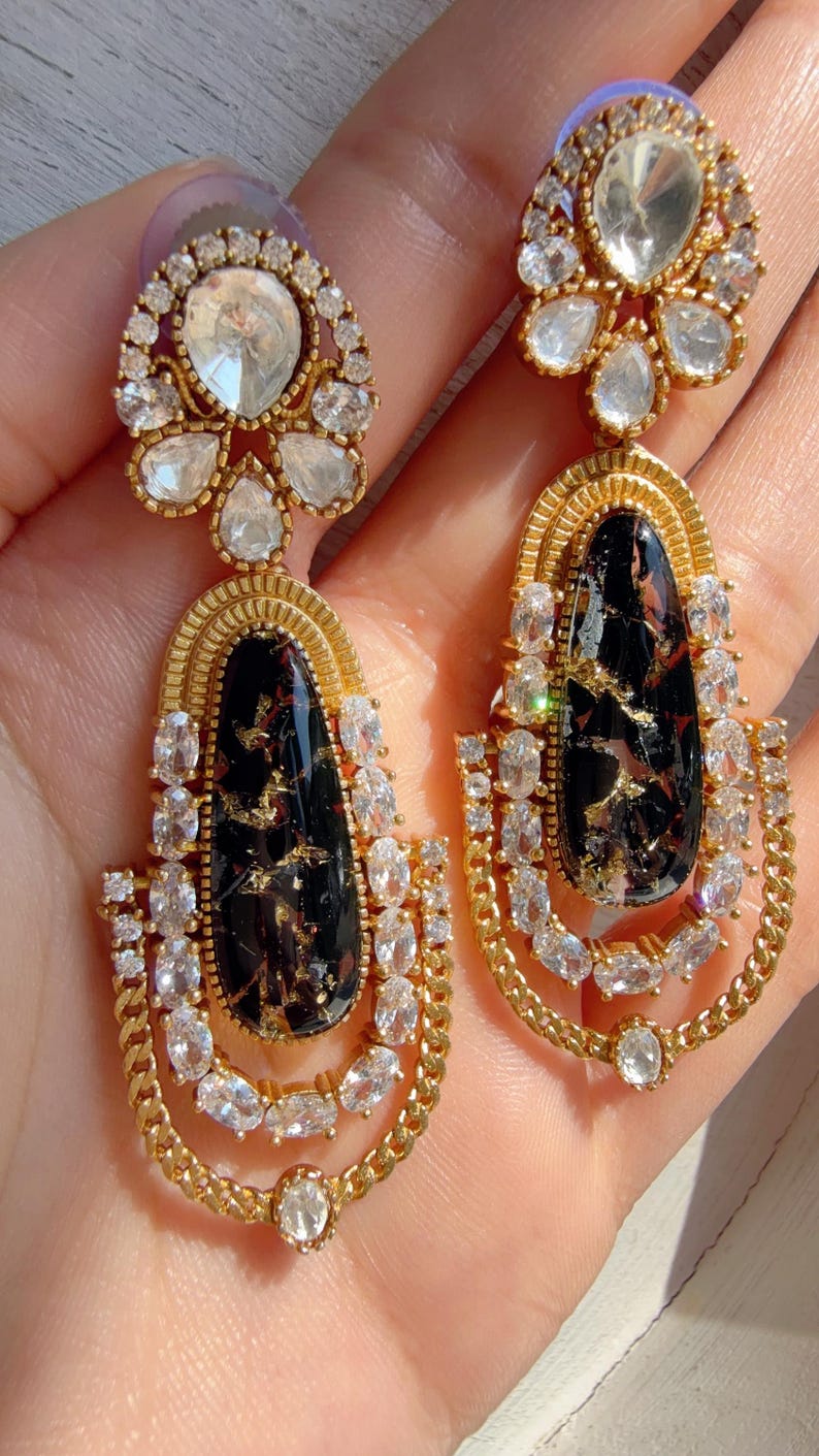 May include: Gold-toned drop earrings with a black, marbled center stone and clear, sparkling accents. The earrings feature a chain-like design and are set with multiple clear crystals. The top of the earrings have a large, clear stone surrounded by smaller crystals.