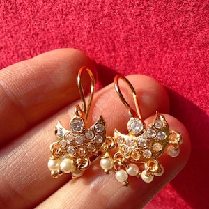 May include: Gold-toned earrings with a bird-like design, adorned with clear crystals and small, dangling pearls. The earrings have a hook closure and are displayed against a red background, showcasing the jewelry's intricate details.
