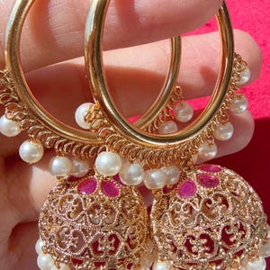May include: Gold-toned hoop earrings with intricate filigree detailing and dangling faux pearls. The earrings feature a gold hoop design with a decorative element and a cluster of pearls. The earrings have a pink gemstone in the center.