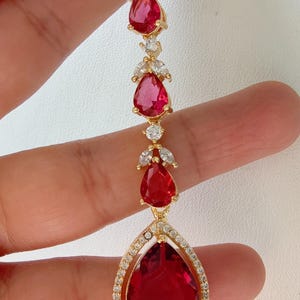 May include: A gold necklace featuring a teardrop-shaped red gemstone pendant surrounded by smaller clear gemstones. The pendant is attached to a chain with smaller red gemstones and clear gemstones.