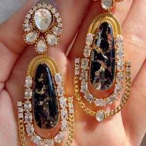 May include: A pair of gold-toned earrings with a black and gold marbled center stone, surrounded by clear crystals. The earrings have a floral-shaped top and a chain-like design at the bottom.