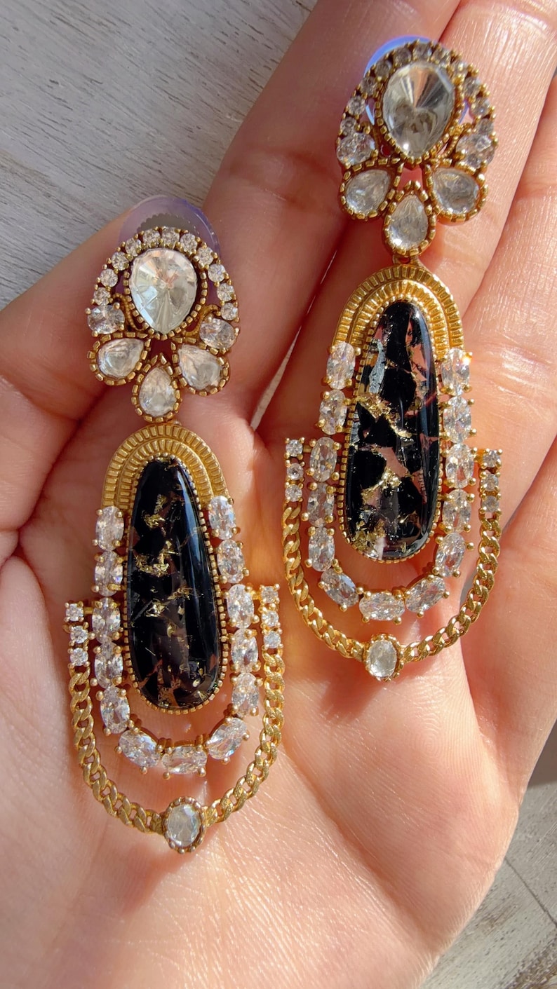 May include: A pair of gold-toned earrings with a black and gold marbled center stone, surrounded by clear crystals. The earrings have a floral-shaped top and a chain-like design at the bottom.