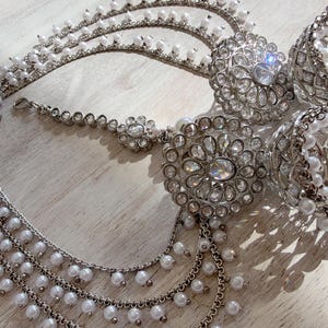 May include: A silver-toned jewelry set featuring two bell-shaped jhumka earrings with intricate metalwork and white faux pearls, connected by a multi-layered chain strand adorned with small white pearls and a central floral-shaped decorative piece.