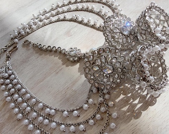 May include: A silver-toned jewelry set featuring two bell-shaped jhumka earrings with intricate metalwork and white faux pearls, connected by a multi-layered chain strand adorned with small white pearls and a central floral-shaped decorative piece.