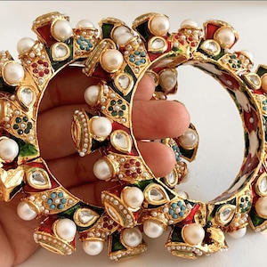 May include: Two gold bangles with intricate designs. The bangles are decorated with red, green, and blue enamel, and are adorned with white pearls and clear stones.