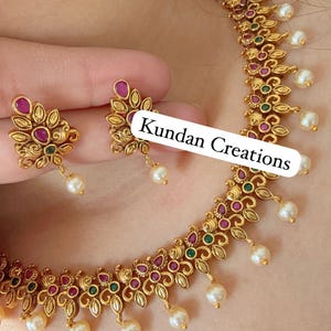 May include: Gold-coloured necklace and earring set with floral and leaf designs, featuring red and green gemstones and dangling pearls. The text "Kundan Creations" is visible on the image.
