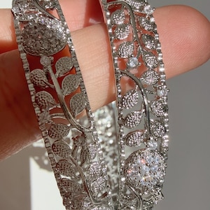 Puede incluir: Dos brazaletes plateados con un diseño de hojas y flores. Los brazaletes están adornados con piedras de circonita cúbica transparentes.