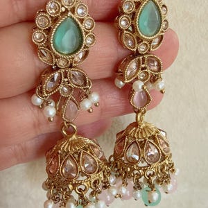 Pode incluir: Brincos dourados com pedras verdes e rosa e p&eacute;rolas. Os brincos t&ecirc;m a forma de uma l&aacute;grima com um charme de sino pendurado.