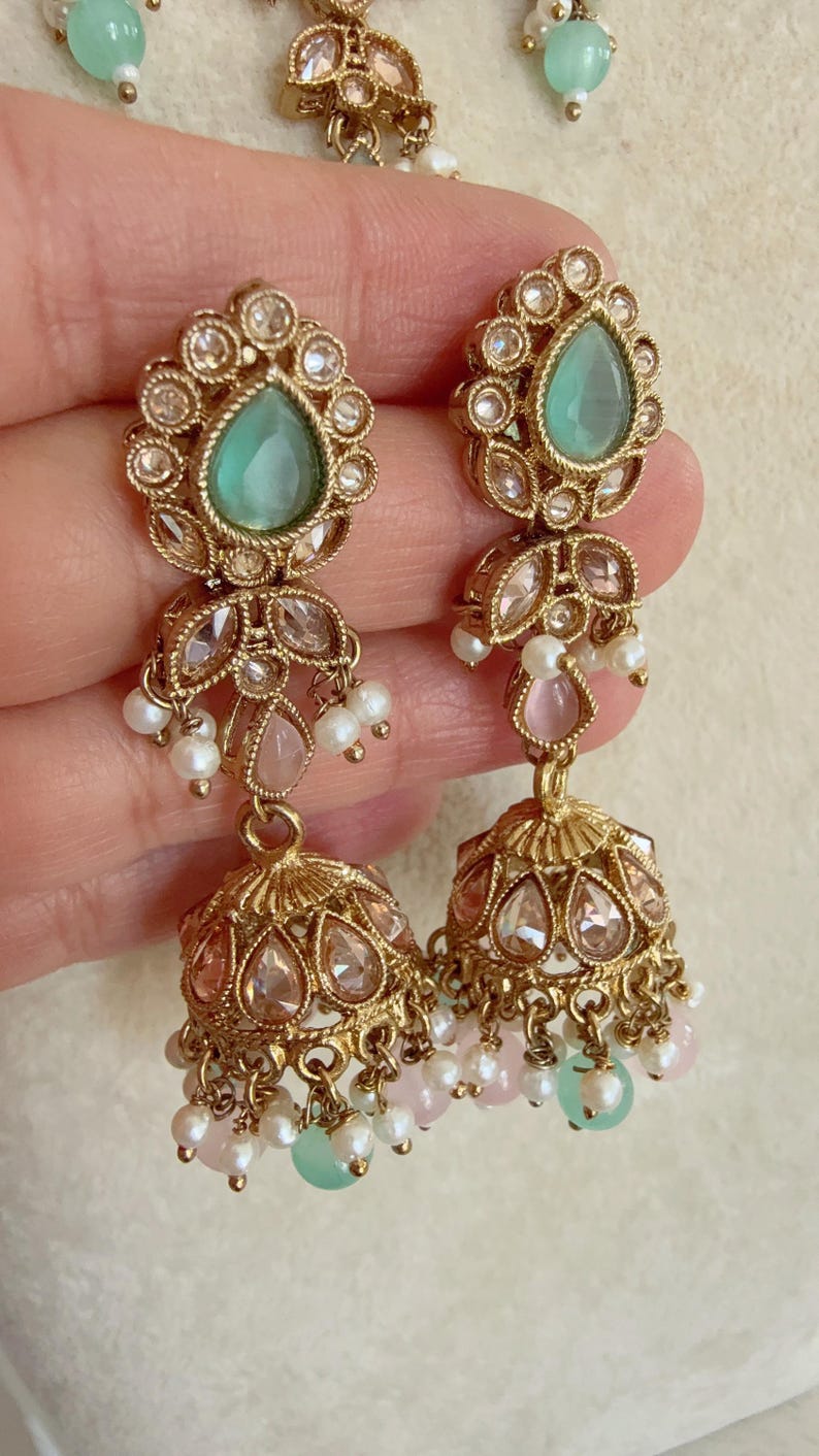 Pode incluir: Brincos dourados com pedras verdes e rosa e p&eacute;rolas. Os brincos t&ecirc;m a forma de uma l&aacute;grima com um charme de sino pendurado.