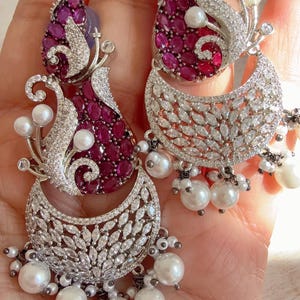 Puede incluir: Elegantes pendientes plateados con diseño de media luna, adornados con cristales transparentes brillantes, piedras preciosas de color rojo intenso y perlas blancas colgantes. Los pendientes tienen un diseño floral y de hojas, perfectos para ocasiones especiales.