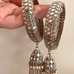 May include: A pair of gold bangles with mirrored detailing and pearl tassel charms. The bangles are decorated with small, round mirrors.