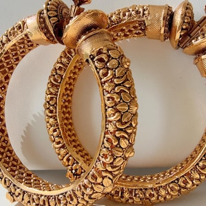 May include: Two gold-tone bangles with intricate floral designs. The bangles are thick and have a rounded shape.