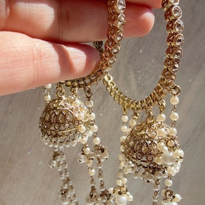 May include: Gold-toned hoop earrings with intricate detailing and dangling pearl accents. The earrings feature a series of clear, round embellishments along the hoop and a bell-shaped pendant. The design is ornate and elegant, suitable for special occasions.