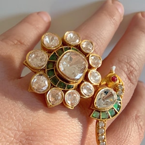Könnte beinhalten: Goldfarbener Ring mit einem großen, klaren Edelstein, umgeben von kleineren Steinen in einem floralen Design. Ein zweiter Ring, geformt wie ein Vogel, hat einen klaren Edelstein, grüne Akzente und ein rotes Auge. Der Ring befindet sich an einer Hand.