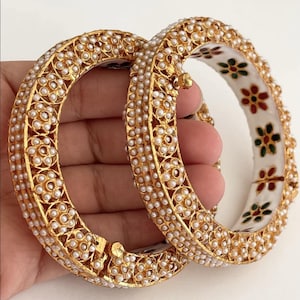 May include: Two gold bangles with white pearl accents. The bangles are decorated with intricate floral designs and have a white enamel base with colorful floral patterns.