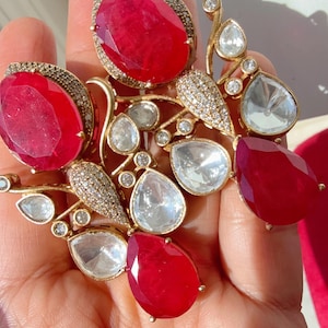 May include: A gold-toned jewelry piece featuring large, oval and teardrop-shaped red gemstones, interspersed with clear, pear-shaped stones and small, sparkling accents. The design has an organic, floral-like arrangement.