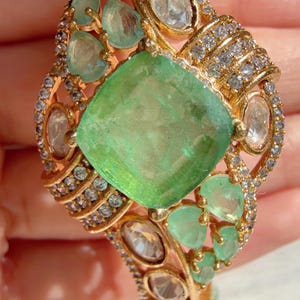 Puede incluir: Un brazalete dorado con una gran gema verde claro de corte cuadrado en el centro. El brazalete está adornado con gemas verdes más pequeñas en forma de corazón y piedras ovaladas transparentes, todo ello en un diseño detallado y ornamentado.