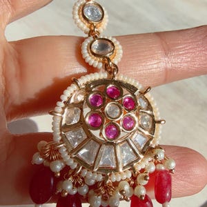 May include: Gold-toned jewelry piece with a central circular design featuring pink and clear gemstones, surrounded by white pearls. The piece has a dangling cluster of red, teardrop-shaped beads and small white pearls.