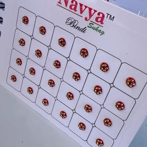 May include: A sheet of Navya Bindi, a traditional Indian forehead decoration. Each bindi is a small, red circle with a decorative, gold-coloured centre. The sheet contains multiple bindis, arranged in a grid pattern.