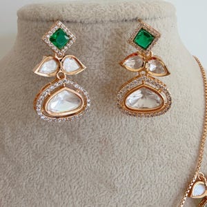 May include: A gold necklace and earrings set with green and clear stones. The necklace has a delicate chain and multiple pendants with a teardrop shape. The earrings are dangle style with a square green stone at the top and a teardrop shaped clear stone at the bottom.