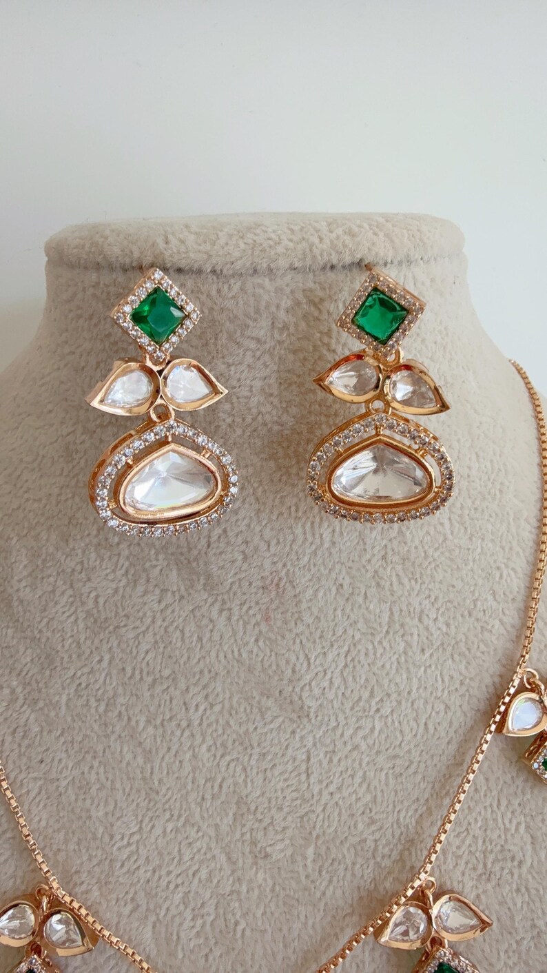 May include: A gold necklace and earrings set with green and clear stones. The necklace has a delicate chain and multiple pendants with a teardrop shape. The earrings are dangle style with a square green stone at the top and a teardrop shaped clear stone at the bottom.