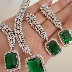 Peut inclure: Un ensemble de bijoux éblouissant comprenant un collier et des boucles d'oreilles. Le collier et les boucles d'oreilles sont sertis d'un métal argenté incrusté de petits cristaux transparents. Chaque pièce présente une grande pierre précieuse rectangulaire vert émeraude.