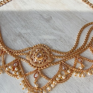May include: A gold-coloured headpiece with multiple strands and delicate pearl accents. The central design features a large, ornate pendant with a clear gemstone. The headpiece is designed with a chain for securing around the head.